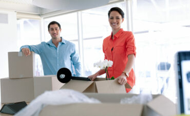 Front view of Caucasian business people looking at camera while unpacking office belongings from cardboard boxes on table in office
